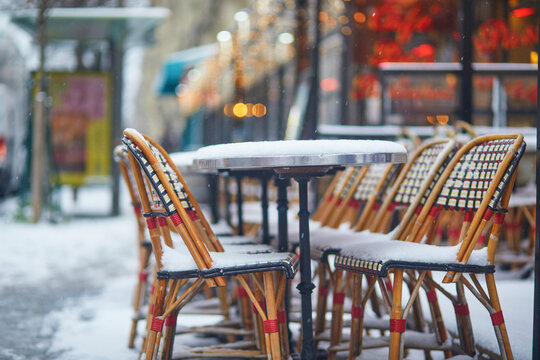 Empty Paris cafe terrace during winter snowfall, with classic bistro chairs and round tables covered in fresh snow