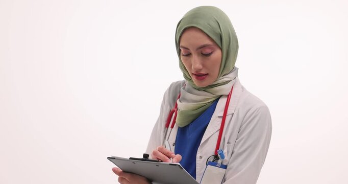Female doctor in hijab writes down symptoms of patient in document on clipboard. Physician creates medical record for future diagnosis at appointment