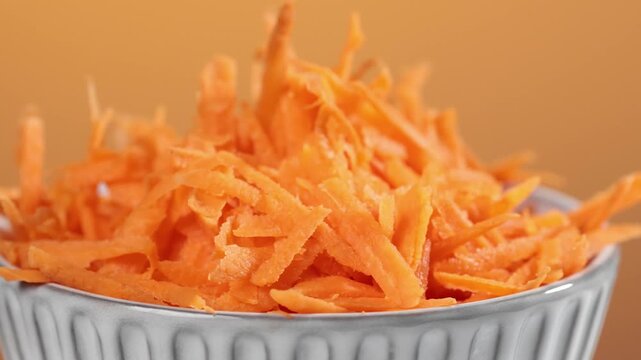 Freshly grated carrots in salad bowl rotate against orange background, closeup of vegetables, preparing healthy dieting vegan meal