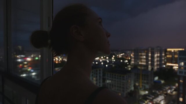 Storm over the city. Woman watches thunder storm with lightnings from the balcony of her apartment on a top floor with city view on the background