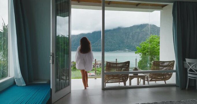 Long-haired tourist woman wakes and walks barefoot onto a private luxury resort balcony, savoring morning sunlight and enjoying panoramic views of Mount Batur, lake, tropical Bali scenery. Back view