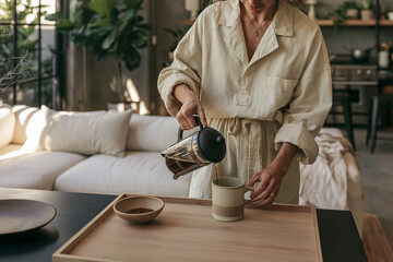 Fototapeta premium Young woman in beige lounge set pouring coffee from French press into ceramic mug in modern loft