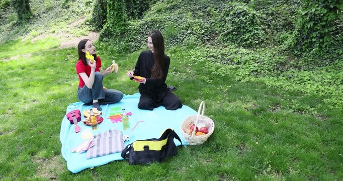Young female friends eat healthy snacks as sit on blanket in green park. Women maintain small picnic with food and ukulele on shady lawn grass