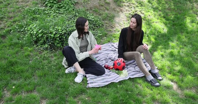 Happy female friends share soda drink as sit on picnic blanket in green park. Woman gives paper cup and opens bottle with companion on shady lawn