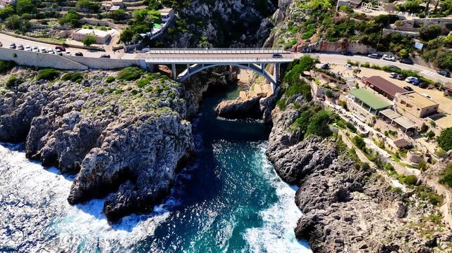 Aerial drone zoom out revealing Ciolo Bridge spanning a rocky coastal canyon above a deep blue Mediterranean inlet in Gagliano del Capo, Puglia, Italy, with cars crossing the bridge.