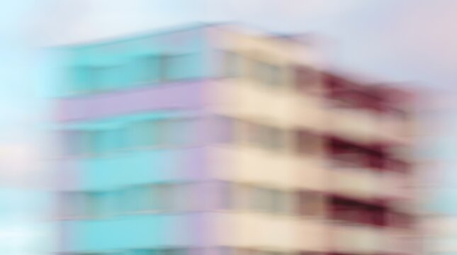 A soft, blurred image of a colorful, modern apartment building under a pastel sky. The building features rows of windows and balconies, with pastel hues of blue, pink, and cream dominating the facade