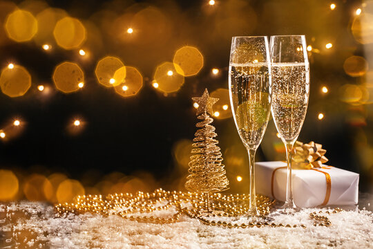 Two champagne flutes beside golden wire Christmas tree and wrapped gift on glittering snowy surface with warm bokeh lights, festive holiday still life arrangement