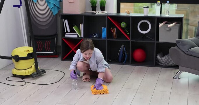 Teenage girl in rubber gloves sprays detergent onto parquet in room. Female teenager cleans floor with rag as sits near rack. Housework routine