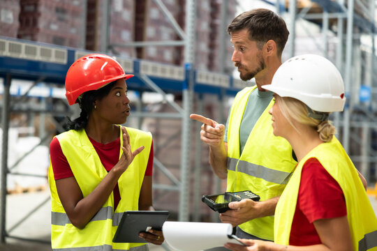 Diverse group of warehouse workers having a serious discussion or argument in a distribution center, wearing safety vests and helmets