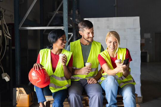 Diverse group of warehouse workers wearing safety vests enjoying a coffee break, laughing, and using a smartphone on a pallet