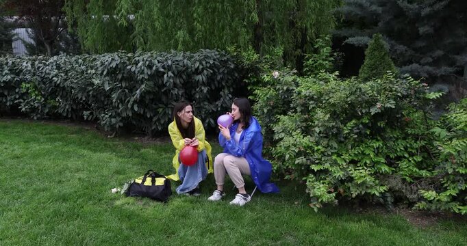 Positive females in waterproof coats blow colorful air balloons in green park. Young women make rainy day brighter with festive details outdoors