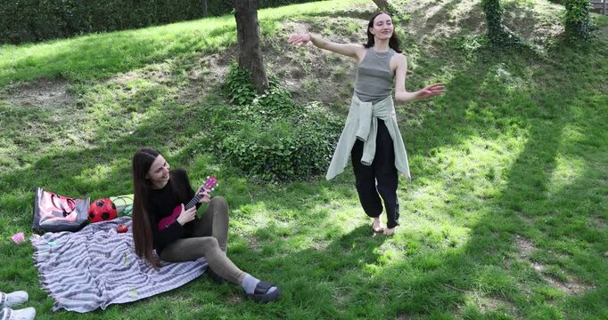 Graceful woman in casual outfit dances as girlfriend plays ukulele sitting in green park. Happy carefree females enjoy music at spring picnic