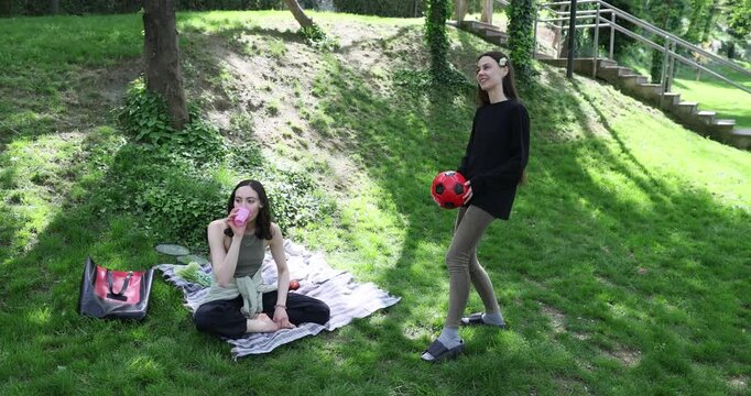 Young woman plays with ball as friend drinks water sitting on blanket in green park. Female girlfriends enjoy outdoor activities at spring picnic
