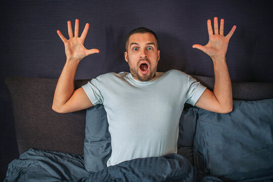 Wondered young man sits on bed and looks on straight on camera. He holds his hands up. Guy is scared and frightened.