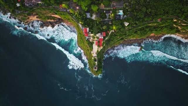 Uluwatu temple and its surrounding green forest sit atop a dramatic cliff, overlooking the powerful waves of the Indian Ocean crashing onto rugged coastline of Bali, Indonesia. Aerial top down shot