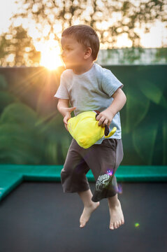 A young boy joyfully jumps on a trampoline in the evening light, holding a bright yellow plush toy. The sun sets behind him, casting a warm glow and creating a playful atmosphere.