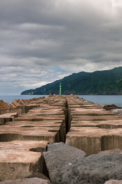 Concrete breakwater leading to a green beacon, protecting the coast of Povoa&ccedil;&atilde;o, S&atilde;o Miguel island, Azores, under an overcast sky with lush green mountains in the background