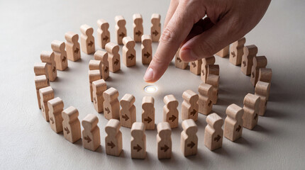 Fototapeta premium Close up of a hand pointing at a glowing center point surrounded by wooden human figures with arrows pointing inward, representing leadership, team goal, target audience, or human resources concept.