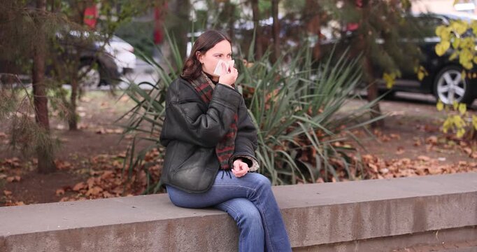 Sick woman uses soft paper tissue to wipe runny nose in autumn. Stylishly dressed female sits on cold concrete parapet in busy city. Slow motion