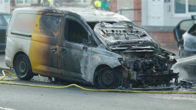 Burned vehicle with open hood and exposed engine compartment after fire. Water spraying toward charred van during firefighting response on urban roadway. Fire damaged commercial van with melted front