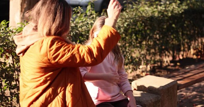 Happy mother and daughter sit together on wooden park bench. Mom and child exchange playful high fives and share warm embrace at sunset. Slow motion