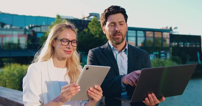 Business man and woman working together outdoors, using a laptop and tablet while looking at the screen and discussing work.