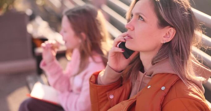 Mother has friendly phone conversation during leisurely walk. Daughter eats sweet refreshing ice cream and admires pleasant surroundings. Slow motion