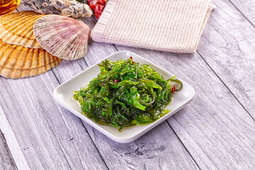 Fresh green seaweed salad with red chili flakes served on white square plate, seashells and textured napkin on weathered wood background © Andrei Starostin