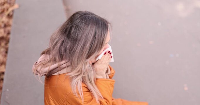 Sick woman sits on concrete parapet and wipes nose with soft tissue. Unwell female in warm autumn jacket wipes runny nose outdoors. Slow motion