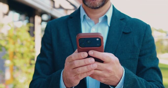 Close-up of an unidentified businessman in a formal suit holding and using a smartphone outdoors.