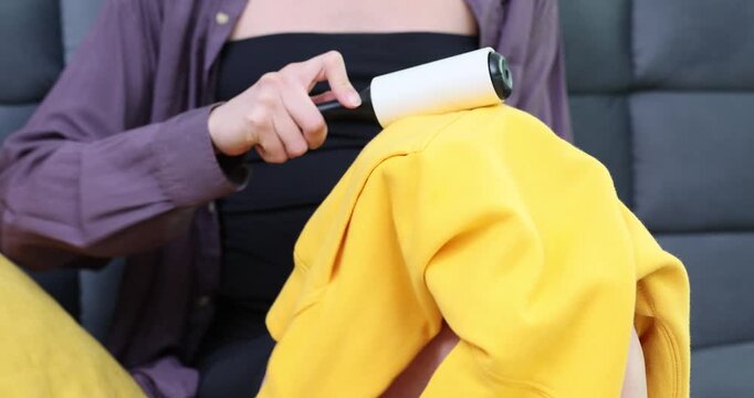 Woman uses sticky lint roller to remove tiny fibers from hoodie. Female sits on sofa and removes delicate fluff and loose fibers time lapse