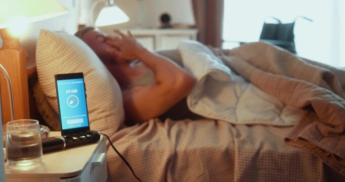 Young man with limited lower-limb mobility wakes to a medication reminder on his smartphone, takes pills from a weekly organizer and drinks water during his morning routine.