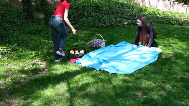 Women spread soft blue blanket across lush green lawn in park. Smiling girlfriends create comfortable picnic area for relaxing afternoon outdoors