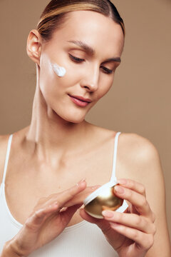 Young woman applies moisturizing cream on face with fingers, has smooth skin and natural makeup, isolated on beige background with soft light.