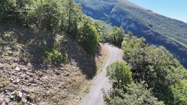 Una strada asfaltata di montagna si snoda tra ripidi pendii rocciosi e alberi fitti sotto un cielo limpido