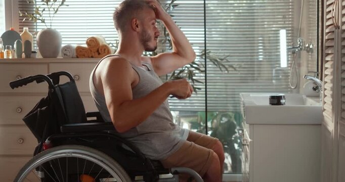Side view of an independent young man in a wheelchair applying hair cream and styling his hair in front of the bathroom mirror during his morning grooming routine.