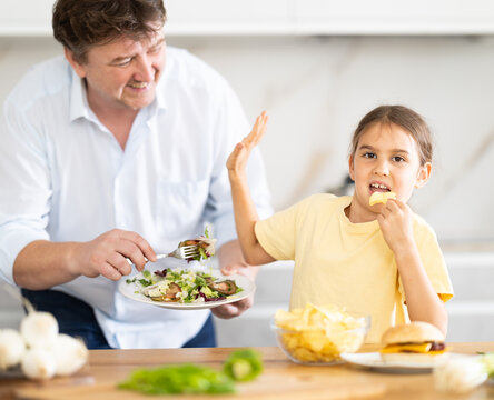 Little daughter eats chips in the kitchen, father offers her healthy vegetable salad