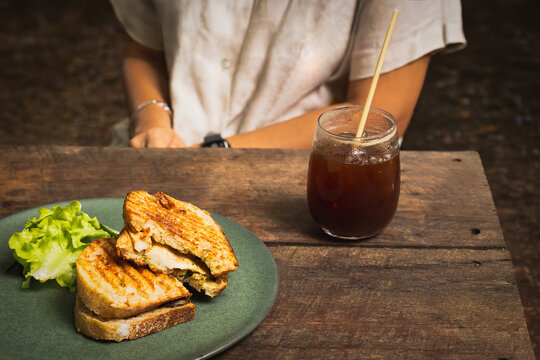 Woman having grilled chicken panini sandwich with glass of iced Americano on wooden table.