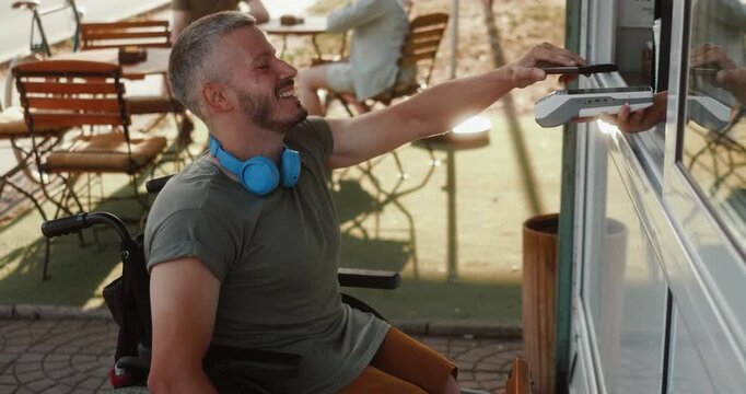 Young man in a wheelchair uses his smartphone for contactless payment at an outdoor cafe, showing independent everyday life, technology use and active social inclusion in the city.
