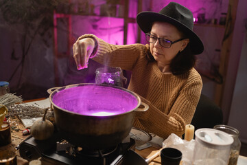 realistic witch brewing a potion in a cauldron. sits at table, adding dried herbs in cauldron. Everyday life intertwined with traditional witchcraft, magic and a mystical atmosphere. © Andrii Lysenko