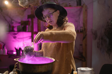 Halloween. Witch in hat adding dried herbs casts spell brews a magic potion in cauldron on fire in scary kitchen. Fantasy. © Andrii Lysenko