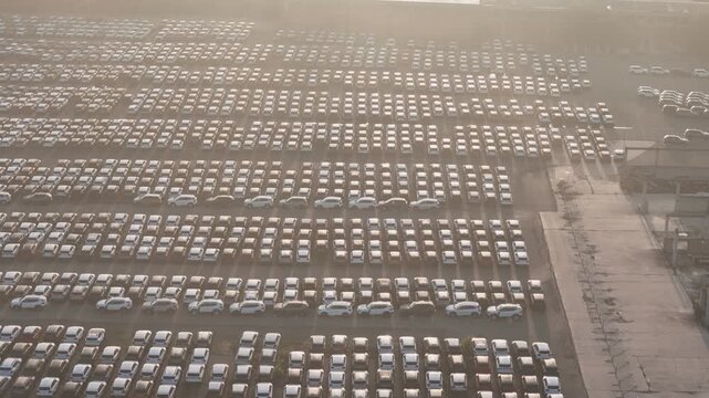 New car storage yard filled with thousands of vehicles arranged in precise rows beside an industrial loading area, illustrating automotive inventory and logistics, aerial shot