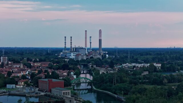 Aerial perspective of a large industrial power plant with towering chimneys standing over a small town crossed by a river at dusk