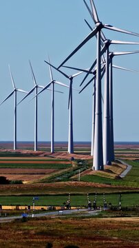 Ascending panning shot of row of spinning wind turbines standing over polder farmland, dike and country road under clear blue sky in Eemshaven Groningen Netherlands