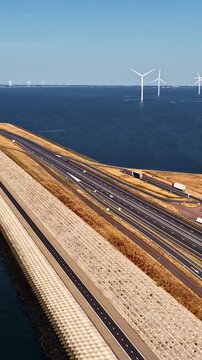Vertical panning shot along A7 highway on Afsluitdijk dike with trucks, service station, small harbor and offshore wind turbines in IJsselmeer near Breezanddijk Netherlands