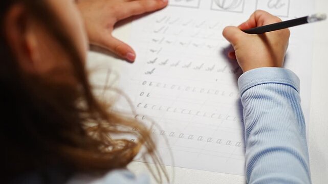 First grade girl practices handwriting with pencil in school workbook. Child learns to write letters during early education with selective focus and shallow depth of field