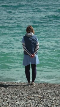 Woman standing alone facing the turquoise sea waves on a pebble beach during a calm afternoon. Static shot.