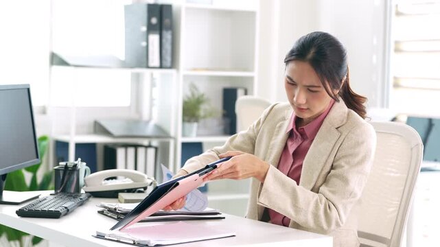 A serious Asian businesswoman is intently searching through a stack of documents while sitting at her desk in her office.
