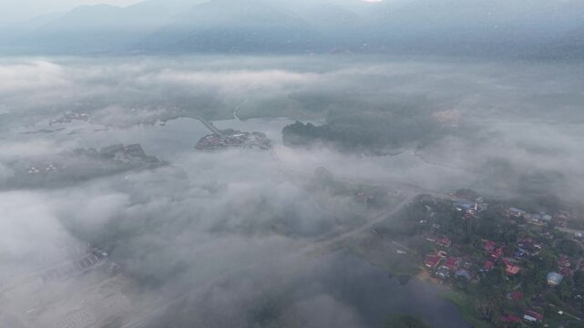 Low clouds cover distant mountains. Valley river flows through red roofed villages at Tasik Raban