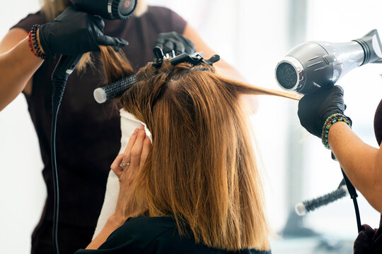 Hairdresser styling client's hair with hair dryer and round brush in beauty salon